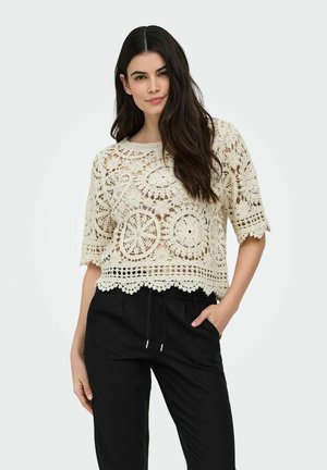 Žena s dlhými tmavými vlasmi, oblečená v krémovom háčkovanom čipkovanom topu a čiernych nohaviciach, pózujúca s jednou rukou v vrecku na jednoduchom pozadí.