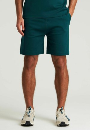 Pantalones cortos de un verde azulado oscuro con una tela texturizada, bolsillos laterales y un corte relajado. Llevados con zapatillas deportivas beige con acentos azules.