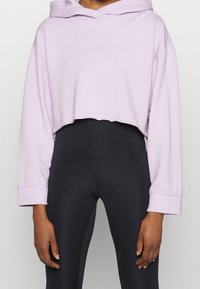 Felpa viola tagliata con cappuccio e maniche lunghe; tessuto morbido; indossata sopra leggings neri a vita alta; cuciture e dettagli minimali.