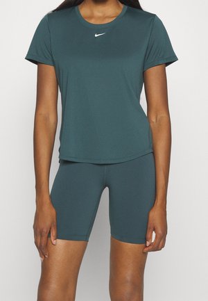 Kvinde iført mørk teal kortærmet sportstop og matchende tætsiddende shorts, stående mod en ensfarvet lys baggrund.