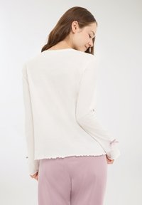 Giovane donna con lunghi capelli castani, indossa una maglietta bianca a maniche lunghe con bordi arricciati e pantaloni rosa, girata leggermente di lato su uno sfondo semplice.