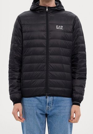 Homme portant une doudoune noire EA7 Emporio Armani avec capuche et un jean bleu clair, debout devant un fond uni.