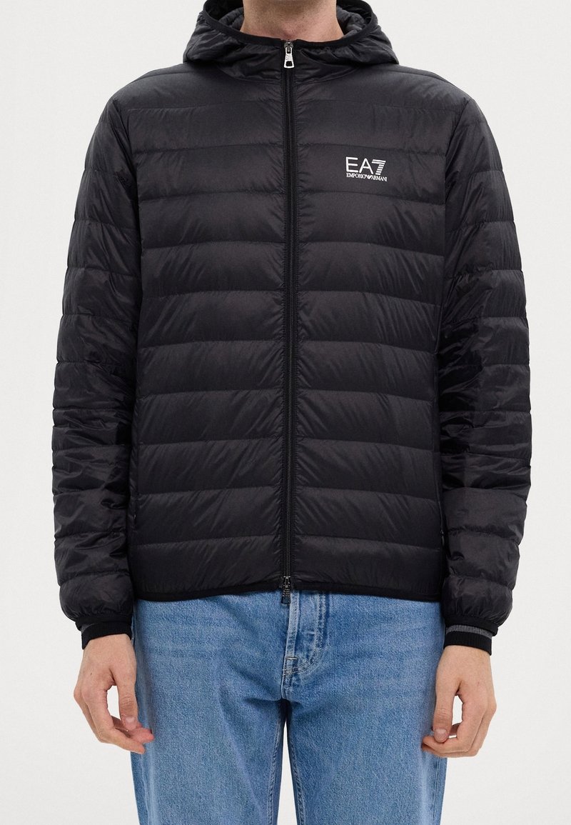 Homme portant une doudoune noire EA7 Emporio Armani avec capuche et un jean bleu clair, debout devant un fond uni.