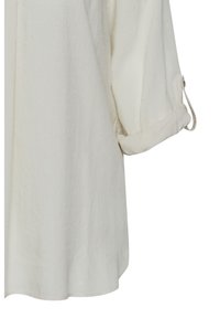 Blouse blanche texturée avec une coupe décontractée, dotée de points embossés subtils et de poignets roulés. Fabriquée en tissu léger.
