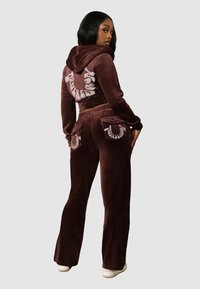 Kobieta ubrana w ciemno-brązowy dres z weluru z kapturem, z krótką kurtką i rozszerzonymi spodniami, pozująca z częściowo odwróconym plecami, widoczne białe trampki.