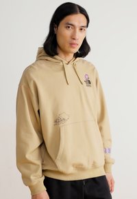Giovane con capelli neri lunghi fino alle spalle, indossa una felpa beige con disegni floreali e un logo, in piedi di fronte a uno sfondo chiaro e semplice.