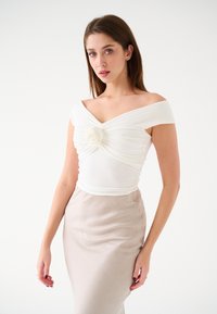 Top bianco scollato sulle spalle con tessuto arricciato e un accento floreale, abbinato a una gonna beige a vita alta. Texture liscia, design aderente.