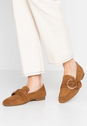 Mocassins en suède marron avec un talon plat, dotés d'une large bandoulière et d'un accent métallique circulaire. Texture lisse et design à bout pointu.