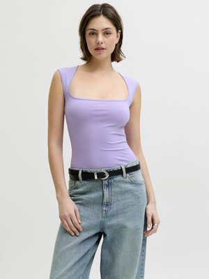 Jonge vrouw draagt een aansluitende lavendelkleurige mouwloze top en lichtblauwe jeans met hoge taille en een zwarte riem, staand tegen een egale achtergrond.