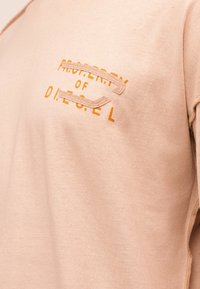 Φωτεινή μπεζ μπλούζα φούτερ με πορτοκαλί κείμενο "PROPERTY OF DIESEL" που είναι κατά το ήμισυ καλυμμένο από ραμμένες υφασμάτινες λωρίδες στην περιοχή του στήθους.