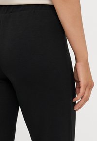 Sorte tights med en glatt tekstur, med høy livvidde og en tettsittende design, sett bakfra med en hånd hvilende på livet.