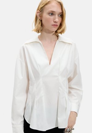 Blondynka w luźnej białej bluzce z głębokim dekoltem w kształcie litery V i długimi rękawami, patrząca na bok na jednolitym tle.