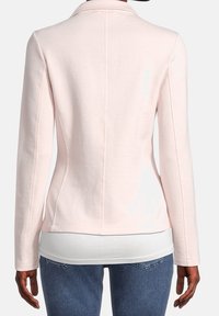 Blazer rose clair en tissu doux, avec une coupe ajustée, un col cranté, des manches longues et des détails de couture au dos. Porté avec un sous-pull blanc.