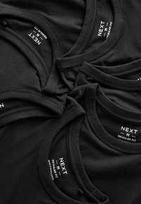 Camisetas de algodón negras apiladas, con cuellos redondos y etiquetas visibles que dicen "NEXT M REGULAR FIT" con el logo. Textura suave y color uniforme.