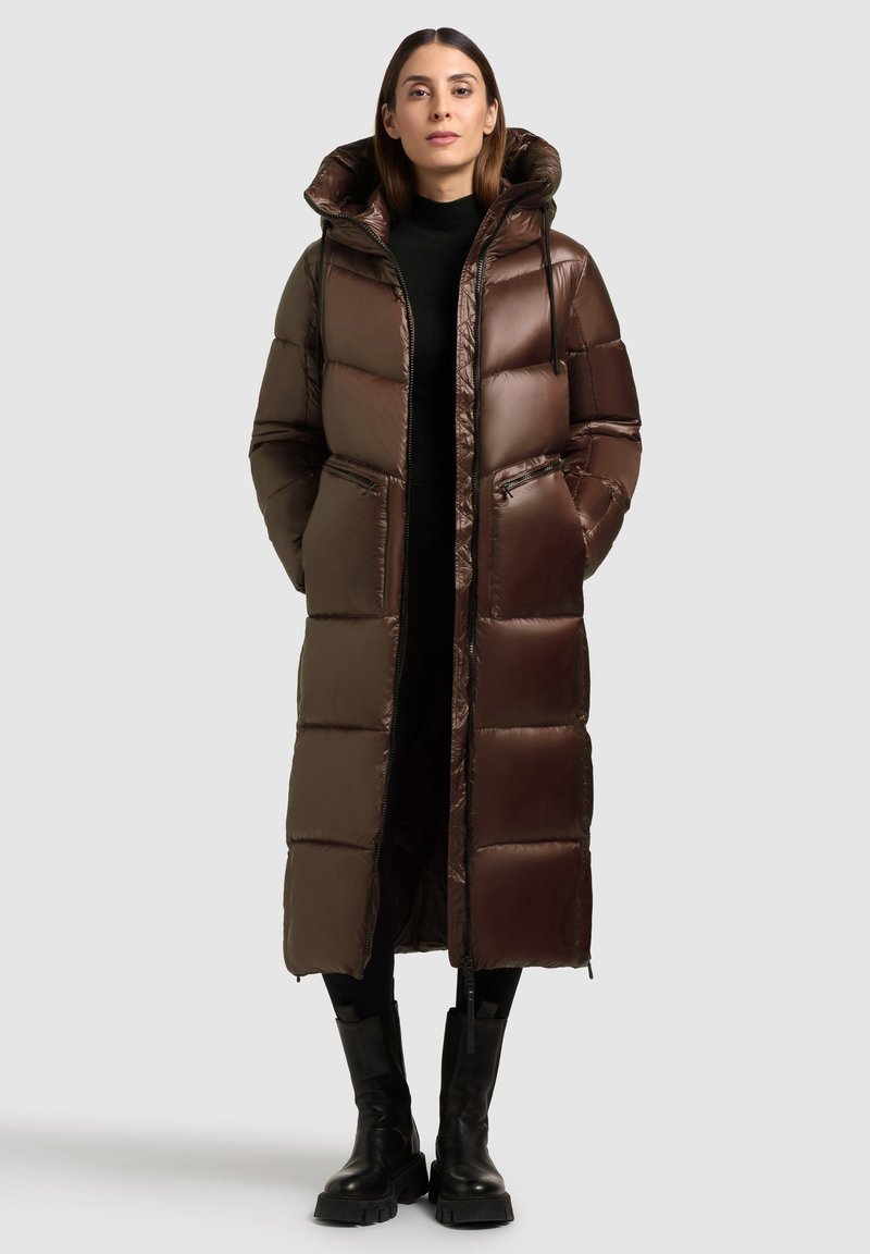 Hosszú, puffos barna tollkabát kapucnival, cipzáras zsebekkel, fényes textúrával és egyenes szabással. Fekete garbó alatt viselt.