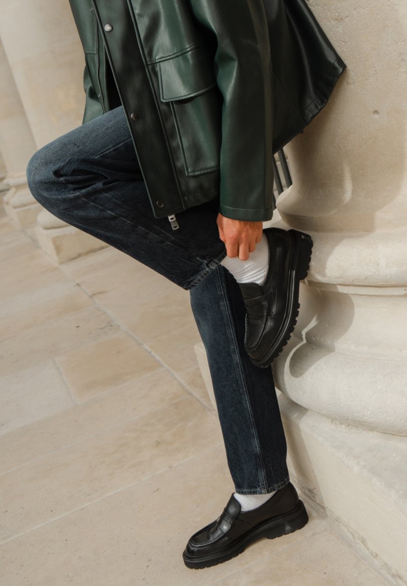 Zwarte leren schoenen met een dikke zool, gedragen met donkere denim jeans en witte sokken, vergezeld van een groene leren jas.
