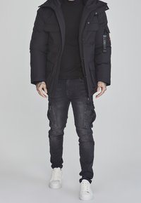 Musta topattu takki, jossa useita etutaskuja, yhdistettynä tummiin farkkuihin, joissa on cargo-taskut, ja valkoisiin tennareihin, neutraalia taustaa vasten.