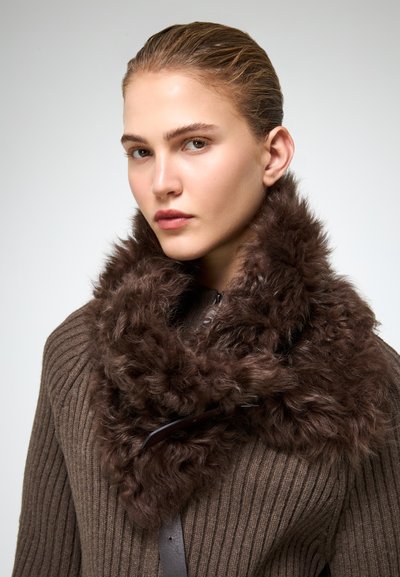 Donna con pelle chiara e capelli tirati indietro, che indossa un maglione marrone a coste e una grande sciarpa di pelliccia marrone scura con una fibbia.