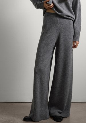 Personne portant un pantalon en maille gris à jambes larges et un pull assorti, se tenant avec une main tenant le pull près de la taille.