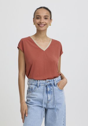 Tânără zâmbitoare, purtând un top cu decolteu în V culoarea portocaliu rugină cu margine bej, blugi cu talie înaltă bleu deschis cu curea de legat, cercei rotunzi aurii, cu mâna în buzunar.