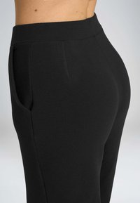 Pantaloni neri con una texture liscia, design aderente, vita alta e tasche laterali. Il tessuto sembra elastico e confortevole.