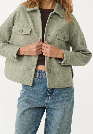 Person iført en salviegrøn knappet jakke med klaplommer over en mørk croptop og højtaljede blå jeans.