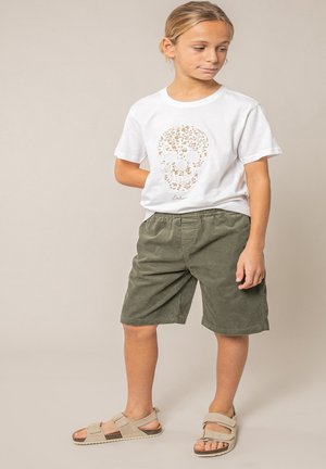 Kind staat, draagt een wit t-shirt met een bloemige schedelprint, olijfgroene korte broek en beige sandalen tegen een neutrale achtergrond.