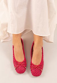 Las bailarinas trenzadas en color rosa presentan una punta redondeada, un acento de lazo decorativo y una superficie texturizada. Los zapatos están combinados con un vestido blanco.