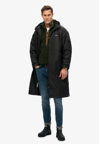 Zwarte waterdichte lange jas met capuchon, gelaagd over een groene fleece, gecombineerd met blauwe spijkerbroeken en zwarte rubberen laarzen. Strak ontwerp, functionele stijl.