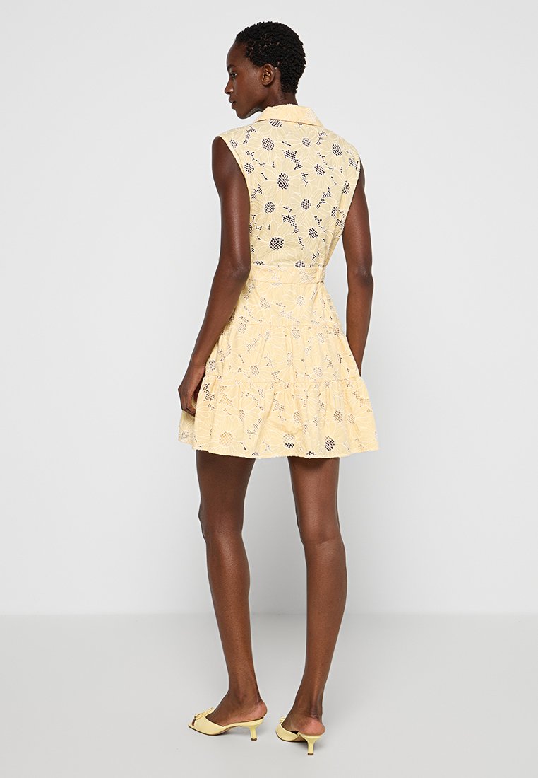 Mujer vestida con un mini vestido amarillo sin mangas con un patrón de encaje floral y mules de tacón bajo a juego, de pie frente a un fondo blanco.