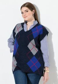 Námořnická modrá bezrukávová vesta s argyle vzorem v šedé, modré a červené barvě. Výstřih do V, jemná textura, vrstvená přes pruhovanou košili.