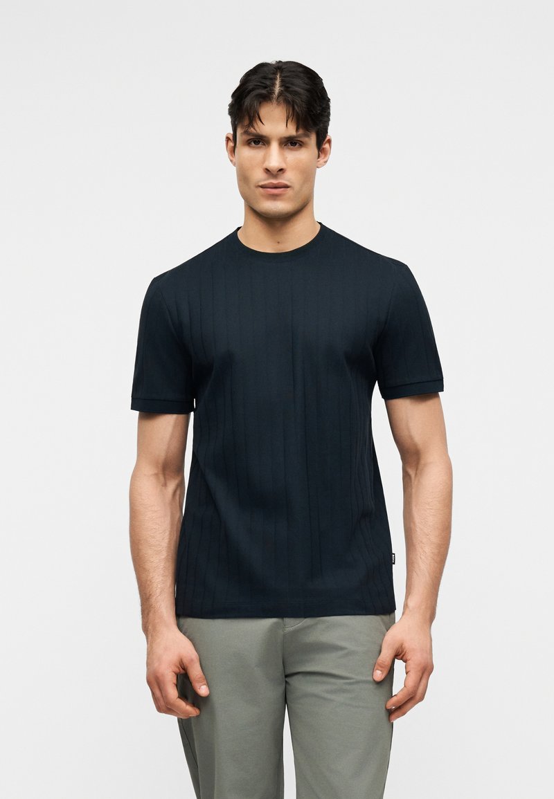 Άνδρας με κοντά σκούρα μαλλιά, που φοράει μαύρο ribbed κοντομάνικο πουκάμισο και ανοιχτό γκρι παντελόνι, στέκεται σε απλό φόντο.