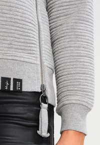 Sweat-shirt gris côtelé avec fermeture éclair. Présente un motif texturé, une poche latérale avec un anneau en métal, et une étiquette en tissu à l'ourlet.