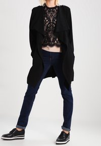 Svart draperad kappa över en svart spets-topp, kombinerad med mörkblå skinny jeans och svarta skor med vita accenter. Slät textur, modern design.