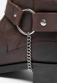 Braunes Lederboot mit einer dekorativen silbernen Kette, die an einem Metallring befestigt ist, mit einem gestapelten Absatz und kontrastierenden Nähdetails.