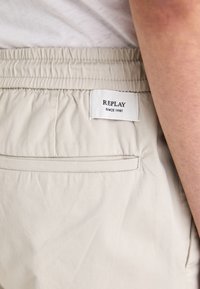 Lys beige shorts med elastisk talje, med en baglomme og et hvidt mærke mærket "REPLAY SINCE 1981." Glat tekstur.