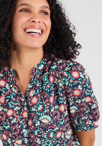 Blusa a maniche corte con motivo floreale su sfondo blu navy, caratterizzata da fiori vivaci in rosa, verde e bianco. Collo plissettato e dettaglio con bottoni.