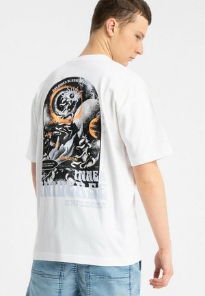 Młody mężczyzna noszący białą, oversize'ową koszulkę z czarno-pomarańczową abstrakcyjną grafiką i tekstem na plecach, w połączeniu z jasnoniebieskimi jeansami.