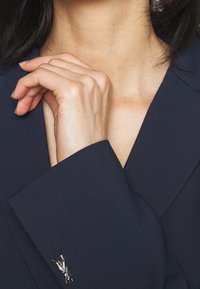 Granatowy blezer o strukturalnym kroju, wykonany z gładkiej tkaniny. Na mankiecie wyróżnia się srebrna broszka w kształcie owada.