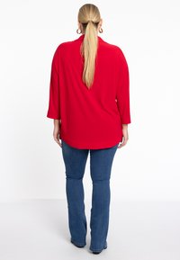 Rotes Langarmshirt mit entspannter Passform und abgerundetem Saum, kombiniert mit blauen Schlagjeans, von hinten präsentiert. Glatte Stofftextur.