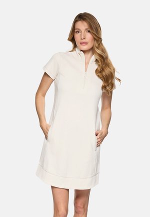 Mujer con cabello largo y ondulado, vestida con un vestido beige de manga corta y largo hasta la rodilla con cremallera frontal y manos en los bolsillos, de pie frente a un fondo liso.