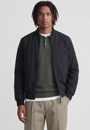 Ung mand med mellemlange dreadlocks iført en sort bomberjakke, grøn tekstureret poloshirt, hvid undertrøje og beige bukser, stående oprejst.
