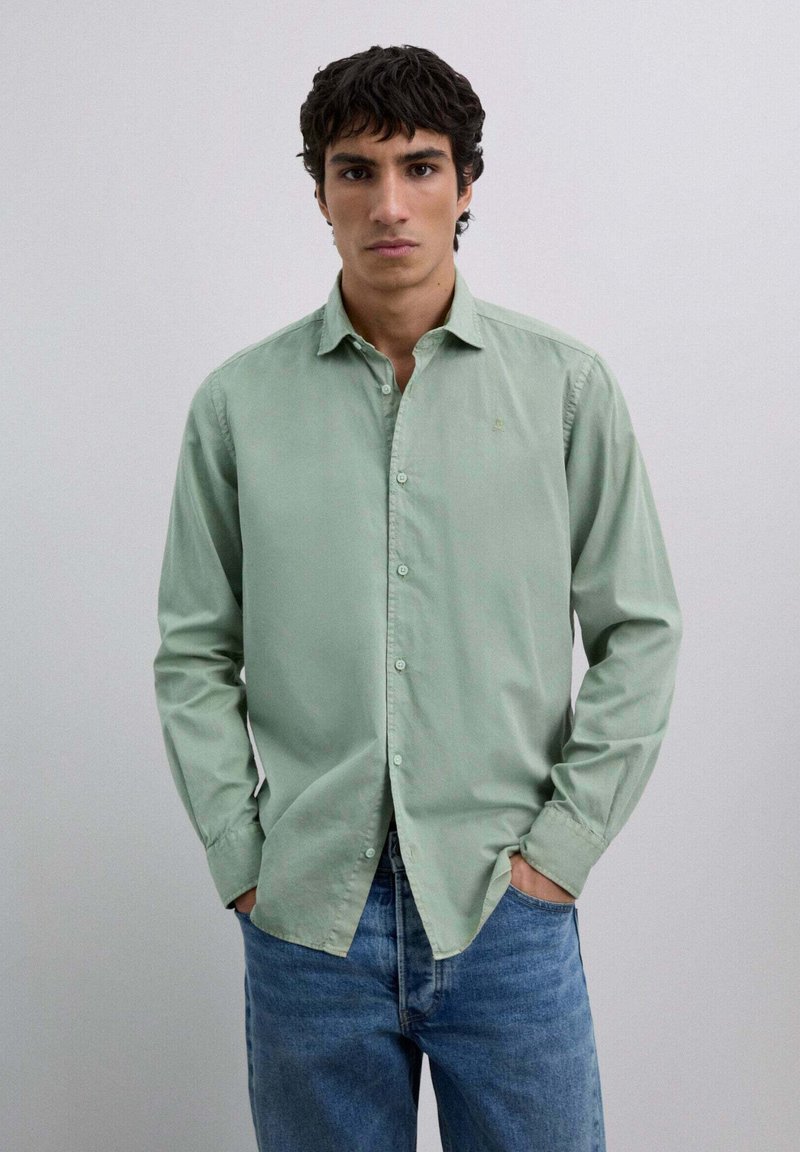 Hombre con cabello oscuro, vistiendo una camisa verde claro con botones y jeans azules, de pie con las manos en los bolsillos contra un fondo claro y sencillo.