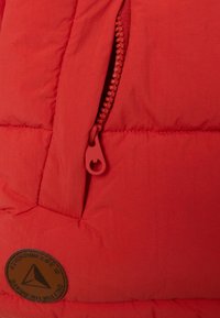 Sezione di giacca a vento rossa con cerniera a contrasto rossa, dotata di imbottitura trapuntata e di un distintivo circolare marrone con testo in rilievo.