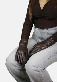 Bruine leren handschoenen met een gladde textuur, voorzien van een aansluitend ontwerp en subtiele accenten. Draagt op handen die rusten op lichtgrijze jeans.