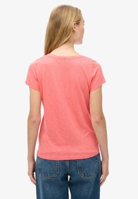 Kvinde med langt blondt hår iført en koralfarvet kortærmet t-shirt og blå denimjeans, set bagfra på en ensartet hvid baggrund.