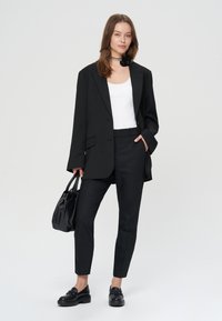 Czarna marynarka na białym prążkowanym topie na ramiączkach, zestawiona z czarnymi eleganckimi spodniami i loafersami. Uzupełniona małą czarną torebką i kwiatowym naszyjnikiem.