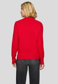 Roter, gestrickter Pullover mit enger Passform, langen Ärmeln und Knopfdetails an den Bündchen, kombiniert mit dunkelgrauen Jeans. Ansicht von hinten.