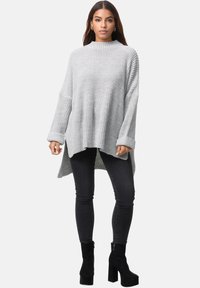 Helles graues, übergroßes Strickpullover mit hohem Kragen, gerippten Bündchen und einem längeren Rückensaum, kombiniert mit schwarzen Skinny-Jeans und Stiefeletten.