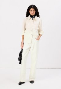 Beige shirt with a collar and front pocket, paired with high-waisted cream pants. Black handbag and shoes with silver accents.